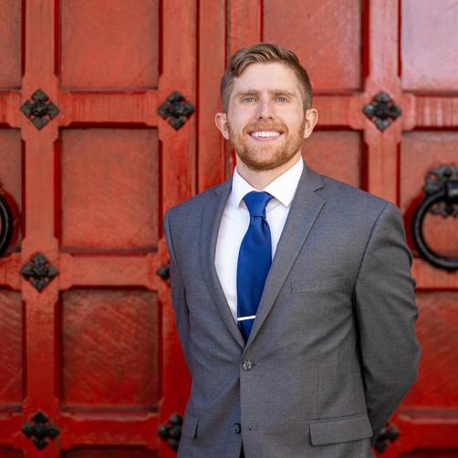 Man in a grey suit and blue tie standing in front of ornate red door.
