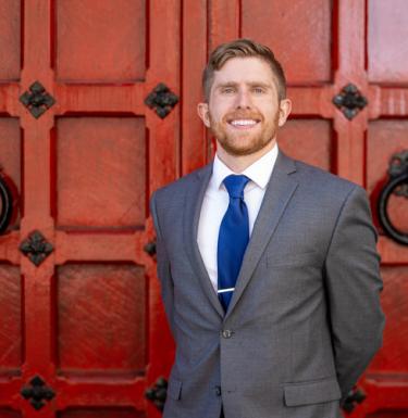 Man in a grey suit and blue tie standing in front of ornate red door.